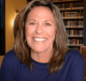 Bright smiling Tracey Devlin with shoulder-length brown hair, wearing a navy blue top, in a cozy library setting.